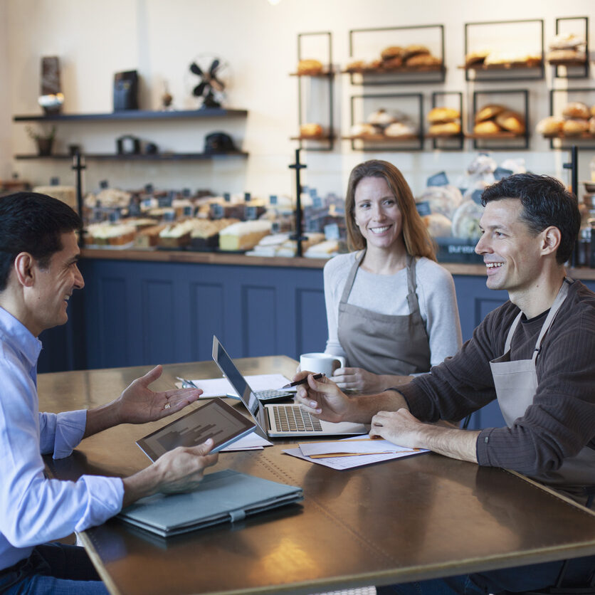 Mature Middle Eastern man financial advisor insurance agent talking advice with mature women and mature man owner staff employee sitting together in small business bakery cafe using laptop computer digital tablet planning
